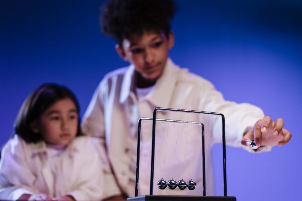 boy pulling on a metal ball of a newton's cradle toy beside a girl